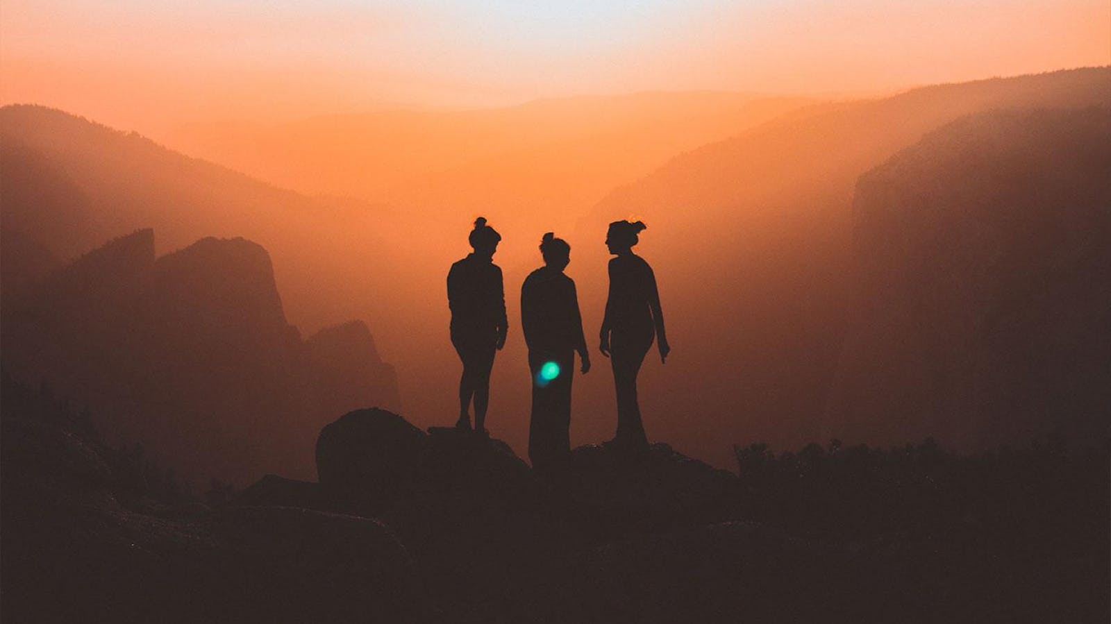 Three women standing on a mountain peak with an orange-tinted sunset view